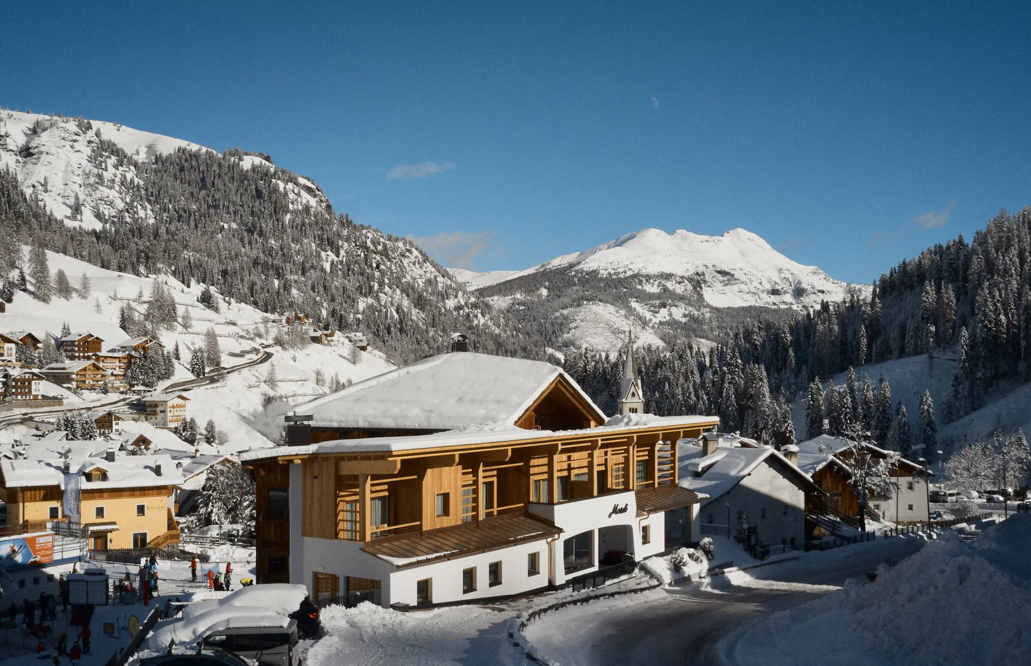 Un pittoresco villaggio innevato nelle Alpi ospita uno chalet con facciata in legno. Montagne innevate si ergono sullo sfondo sotto un cielo azzurro limpido.