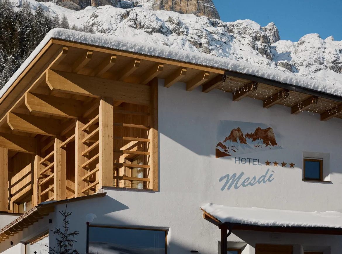 Un hotel alpino in legno con il tetto coperto di neve sorge ai piedi di montagne aspre e innevate sotto un cielo azzurro limpido, trasmettendo un’atmosfera invernale serena.