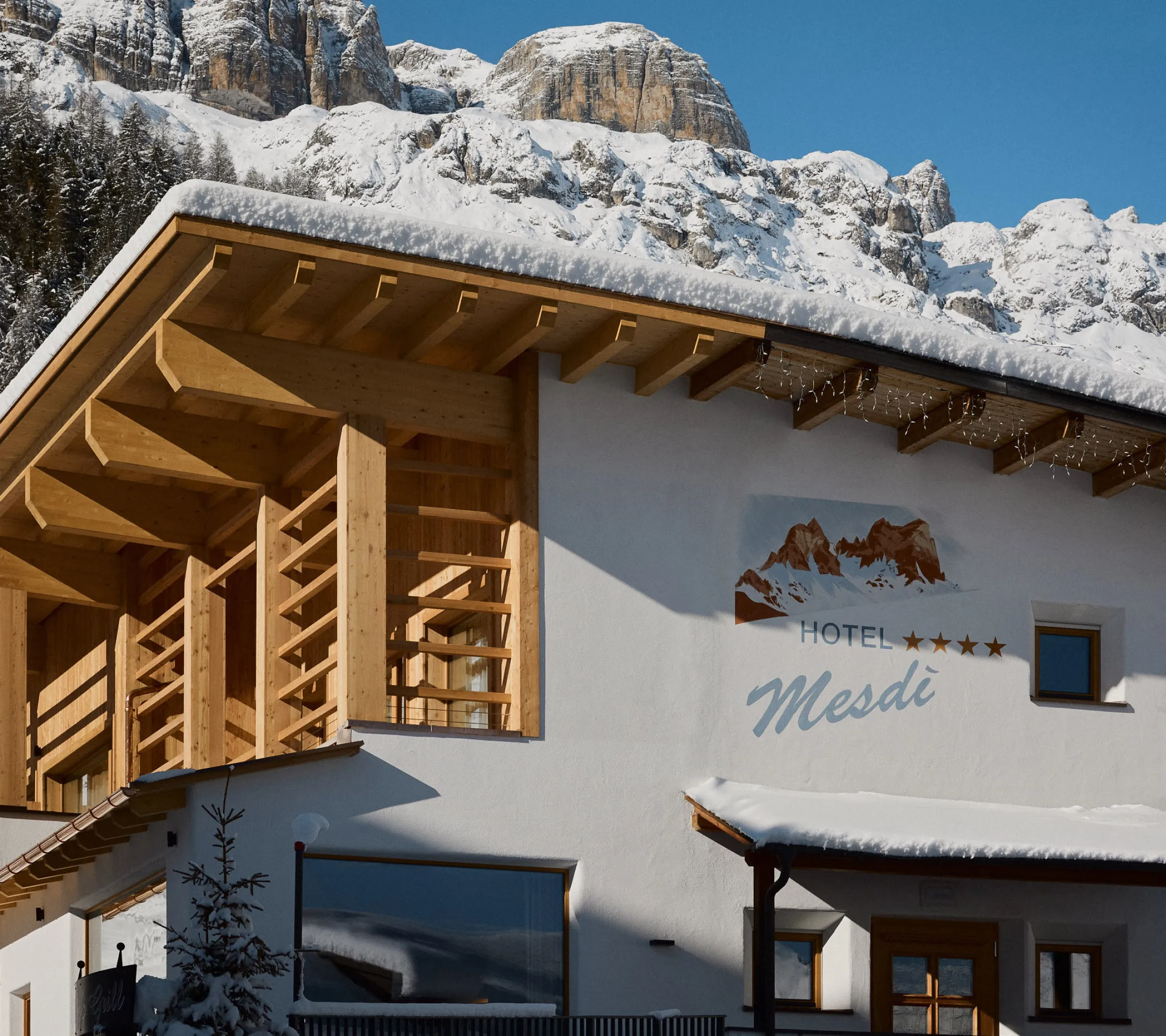 Un hotel alpino in legno con il tetto coperto di neve sorge ai piedi di montagne aspre e innevate sotto un cielo azzurro limpido, trasmettendo un’atmosfera invernale serena.